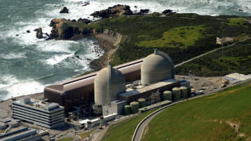 A coastal nuclear power plant with two large concrete domes is situated near rocky cliffs and the ocean. The facility includes multiple buildings, cylindrical tanks, and electrical infrastructure, surrounded by green hills and accessed by a winding road.